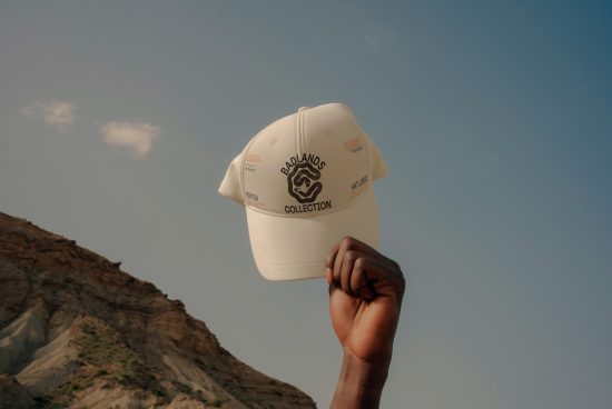 Person holding a white cap with Badlands Collection logo against a blue sky and rocky landscape, ideal for mockup and apparel design templates.